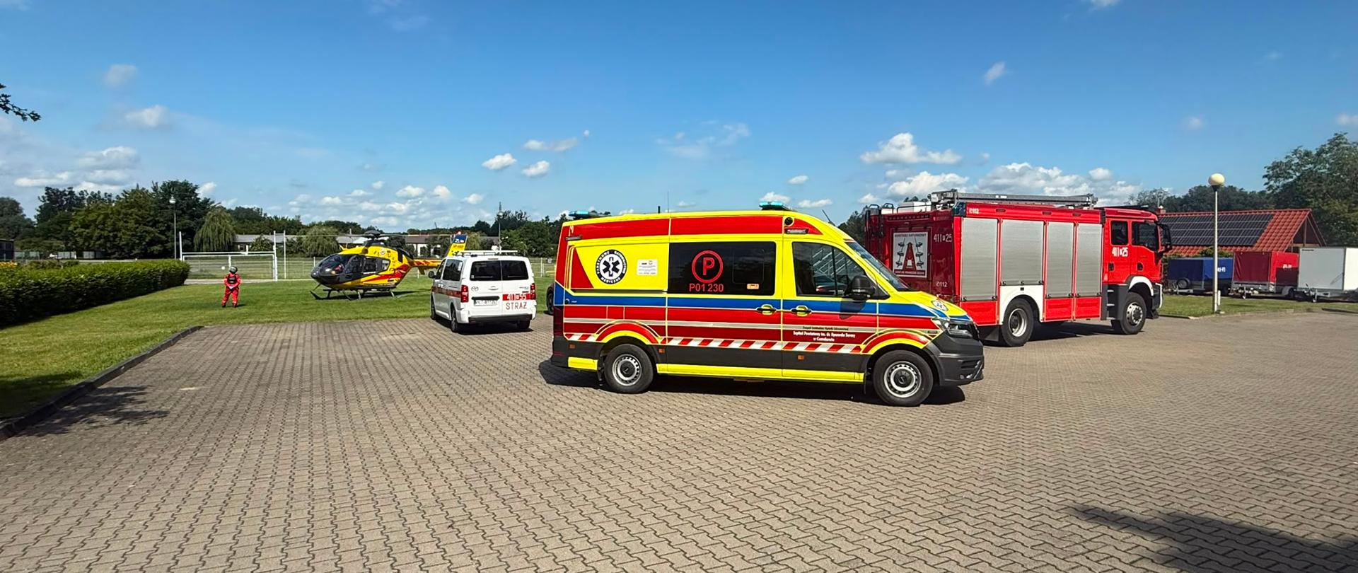 Teren Komendy Powiatowej PSP w Czarnkowie podczas działań ratowniczych. Widoczne są śmigłowiec LPR, ambulans, samochód straży pożarnej i pojazd PSP.