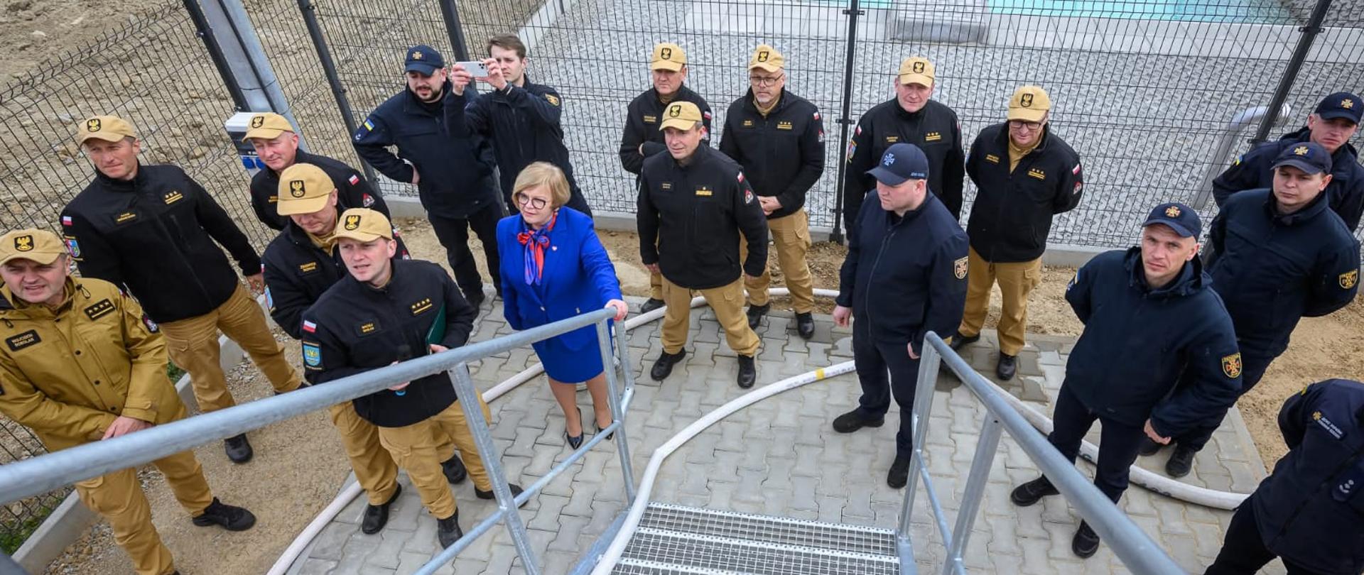 Kolorowa fotografia wykonana w pochmurny dzień na zewnątrz. Przedstawia strażaków polskich w strojach służbowych (piaskowych) z ocieplaczami oraz ukraińskich podczas pokazów przy trenażerze treningowym nowo ustawionym przy komendzie powiatowej PSP w Brzozowie.W tle zbiornik wodny dla płetwonurków.