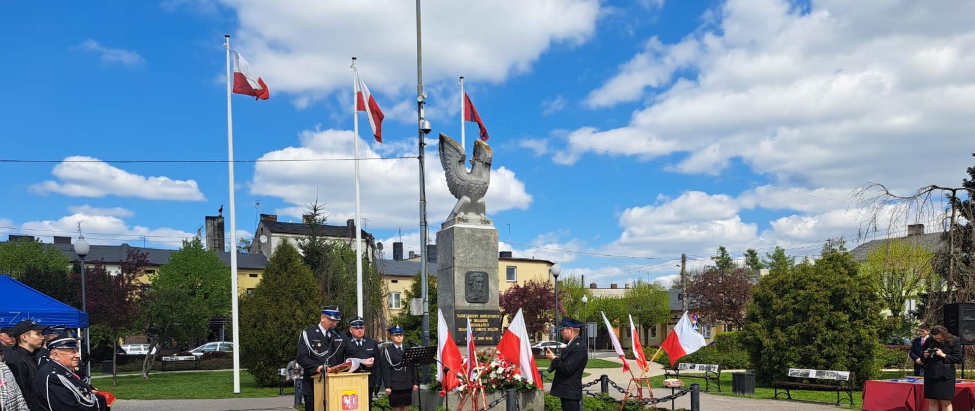 na zdjęciu widzimy pomnik i flagi Polski oraz prowadzącego uroczystość strażaka obok trzech strażaków w ubraniach wyjściowych po lewej stronie widoczni zaproszeni goście w tle drzewa i budynki