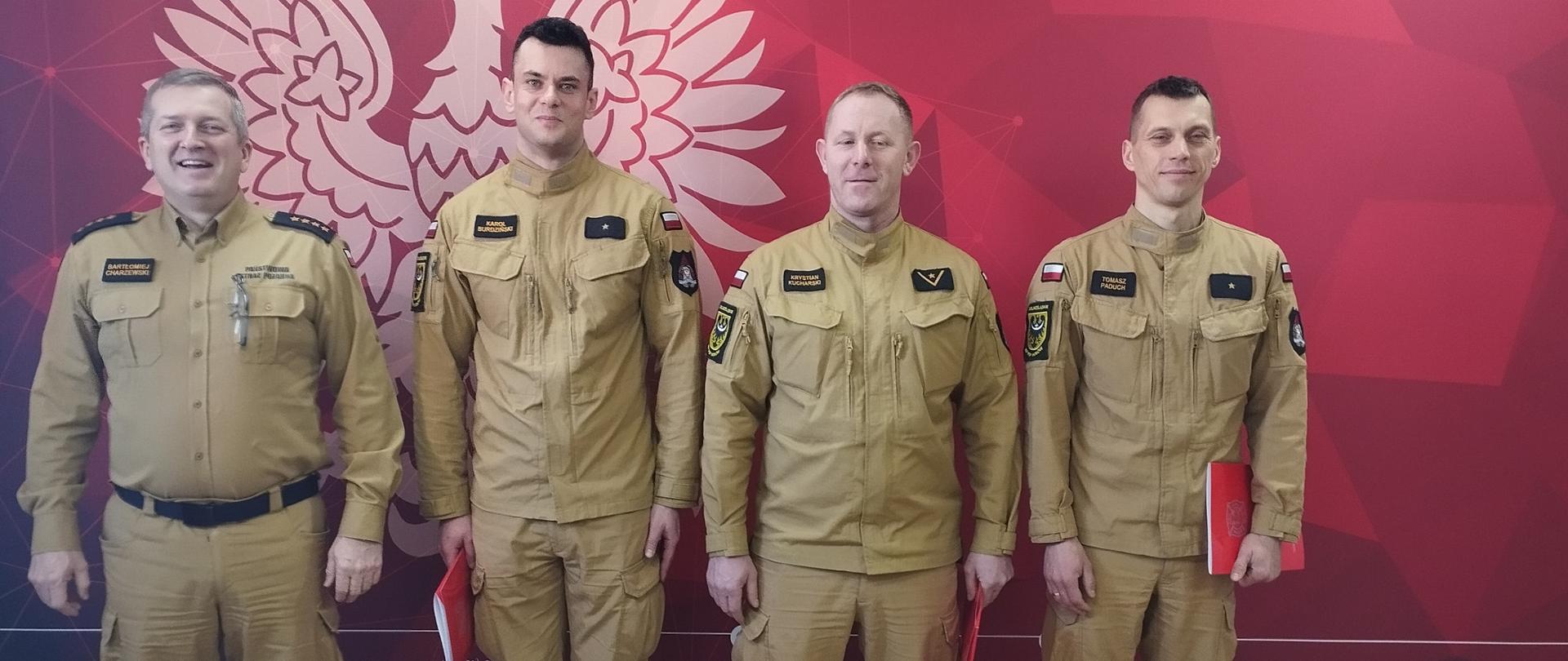 Zdjęcie przedstawia oficjalną fotografię czterech funkcjonariuszy Państwowej Straży Pożarnej, wykonaną wewnątrz budynku na tle pamiątkowej ścianki. Na zdjęciu znajduje się czterech mężczyzn ustawionych obok siebie w jednym rzędzie. Wszyscy mają na sobie oficjalne mundury służbowe w kolorze piaskowym. Mężczyzna po lewej stronie, Komendant Powiatowy PSP w Wołowie ubrany jest w koszulę mundurową z pagonami wskazującymi na stopień starszego kapitana. Pozostali trzej strażacy mają na sobie bluzy mundurowe. Dwóch z nich trzyma w dłoniach czerwone teczki. Wszyscy mężczyźni patrzą prosto w obiektyw i lekko się uśmiechają. Tło stanowi intensywnie czerwona ścianka z białą grafiką. W centralnej części widać duży zarys orła w koronie (godło Polski). U góry widnieje biały napis: „PAŃSTWOWA STRAŻ POŻARNA Komenda Powiatowa PSP w Wołowie”. W prawym górnym rogu znajduje się oficjalne logo Państwowej Straży Pożarnej. Na dole ścianki widoczne są drobne napisy z adresem strony internetowej oraz danymi kontaktowymi komendy.