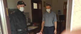  Na zdjęciu widać po lewej stronie strażaka ubranego w czarny mundur typu KDS. Na twarzy biała maseczka. Na głowie czapka z daszkiem. Po prawej stronie osoba duchowna, ubrana w szarą koszulę. Na twarzy biała maseczka. Osoba ta w ręku trzyma opakowanie z maskami ochronnymi koloru białego. Zdjęcie zrobione jest na wejściu do kancelarii. Osoby stoją na czerwonym dywanie. Za osobami widoczne jest biurko. Z tyłu widoczne są drzwi do innych pomieszczeń.
