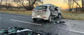 Zniszczony pojazd osobowy stoi w poprzek drogi asfaltowej. Wokół niego widać wiele odłamków plastików i szkła. 