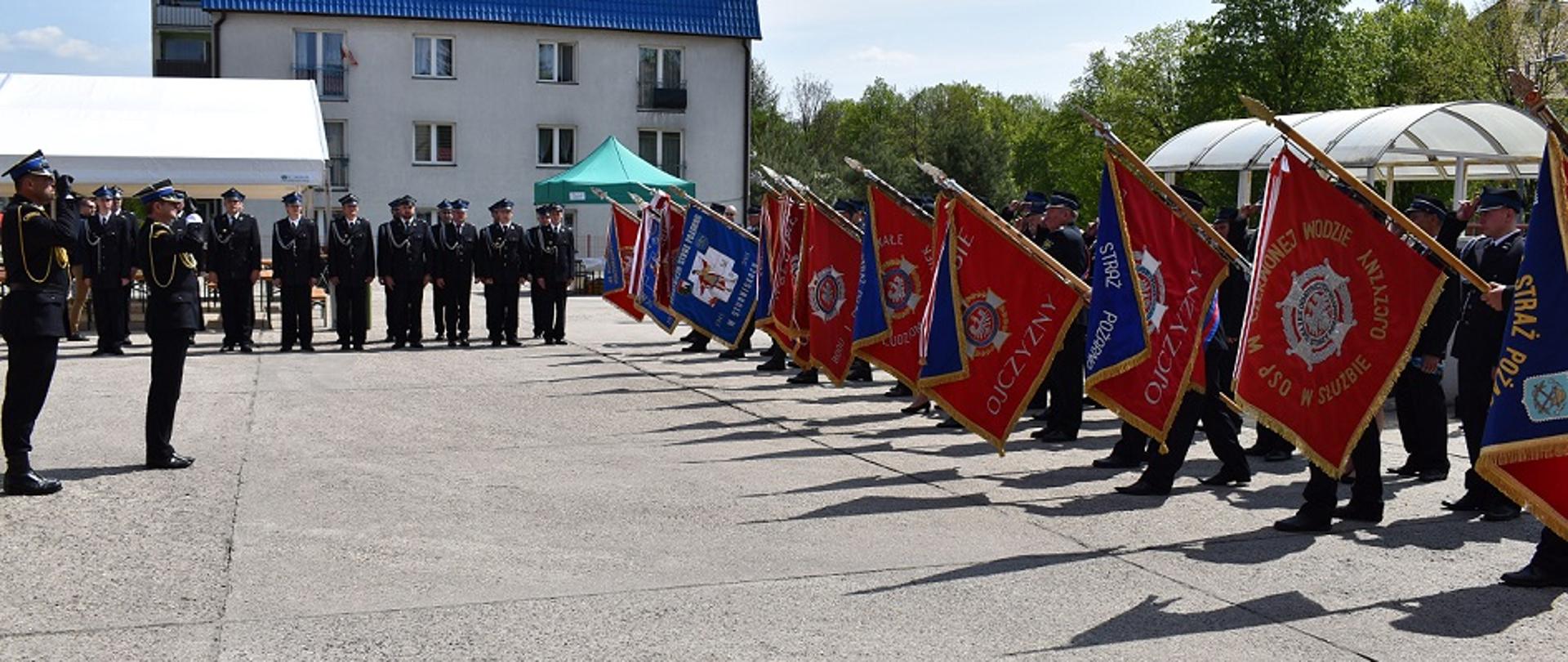 Dzień. Plac z powierzchnią wybetonowaną. W tle zdjęcia widać budynek mieszkalny z niebieskim dachem. Z prawej strony w szeregu stoją poczty sztandarowe, które reprezentują swoje sztandary (sztandar wyciągnięta przed poczet). Z lewej strony zdjęcia dwóch funkcjonariuszy PSP ubranych w mundury wyjściowe w kolorze czarnym, sznur galowy w kolorze żółtym oraz rogatywki oddają honory ( dwa place prawej dłoni przyłożone do daszka rogatywki) zaprezentowanym sztandarom. W tle przed dużym namiotem pokrytym białą plandeką stoi kompania honorowa PSP.