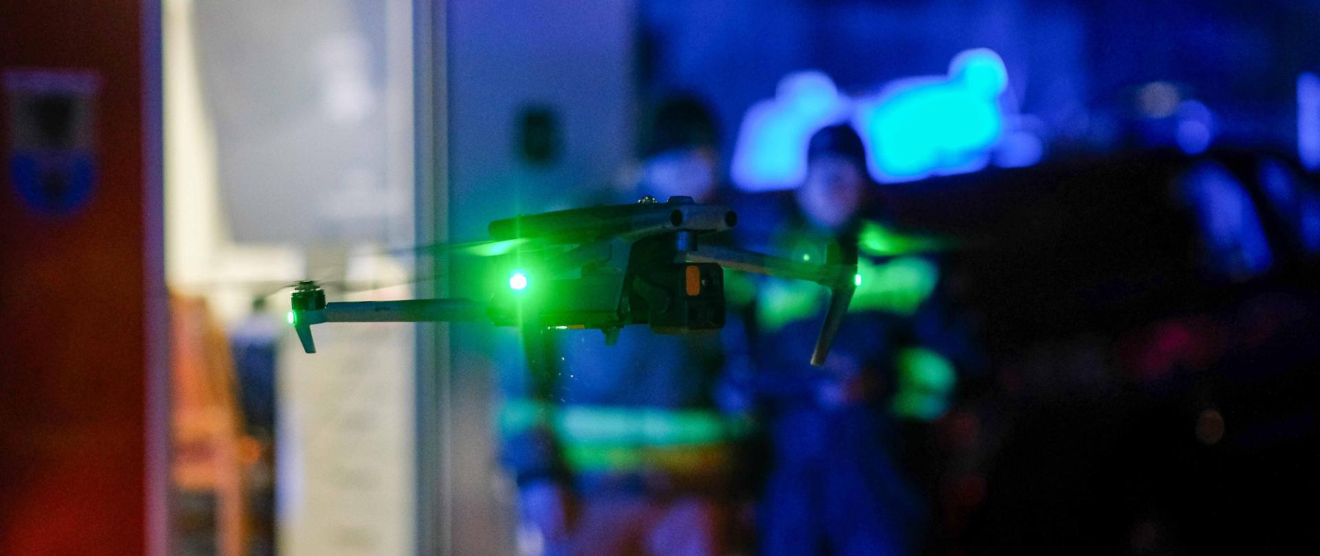 Noc. Na pierwszym panie dron z zielonymi światłami w zawisie. W tle widać sylwetki operatorów drona oraz światła pojazdów uprzywilejowanych.