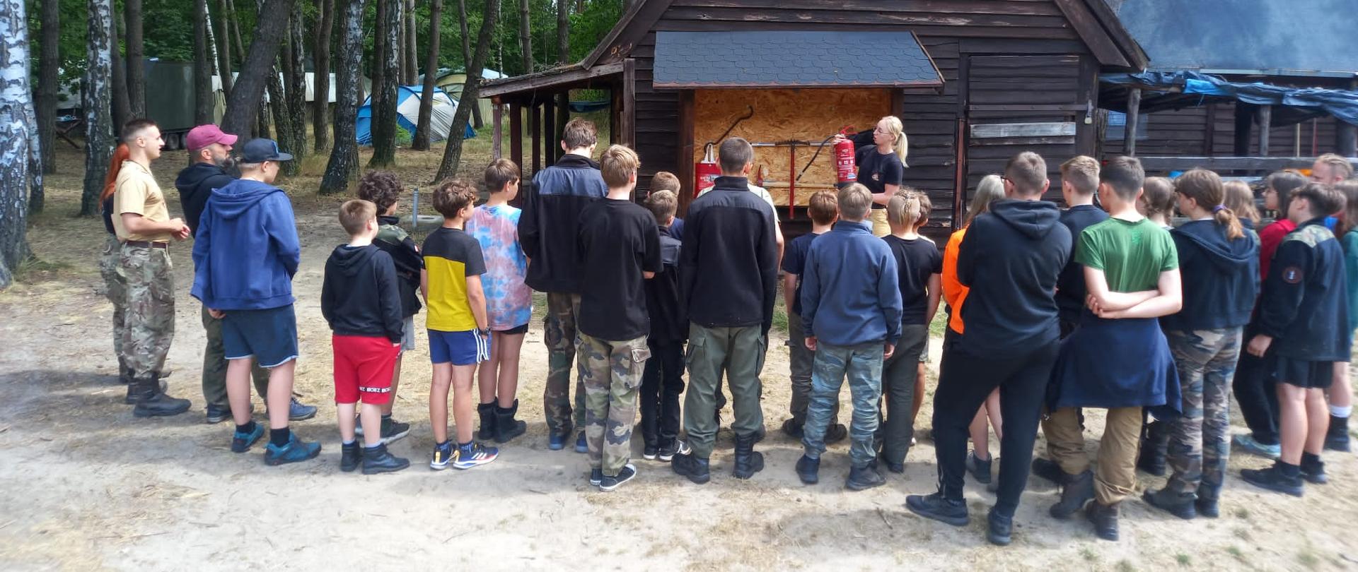 Strażak z gaśnicą na tle drewnianego domu. Przed nim grupa harcerzy