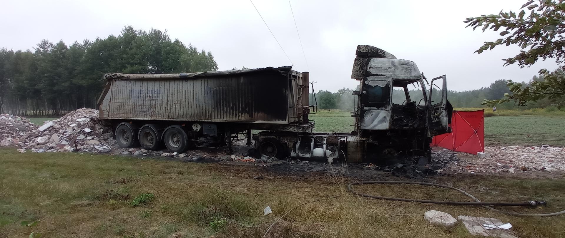 Na zdjęciu widoczny jest wrak spalonej ciężarówki stojącej pod linią średniego napięcia. Naczepa wraz z ciągnikiem stoją na drodze usypanej z gruzu. Drzwi w ciągniku są pootwierane. Po prawej stronie fotografii widoczny jest czerwony strażacki parawan. W tle widać las.