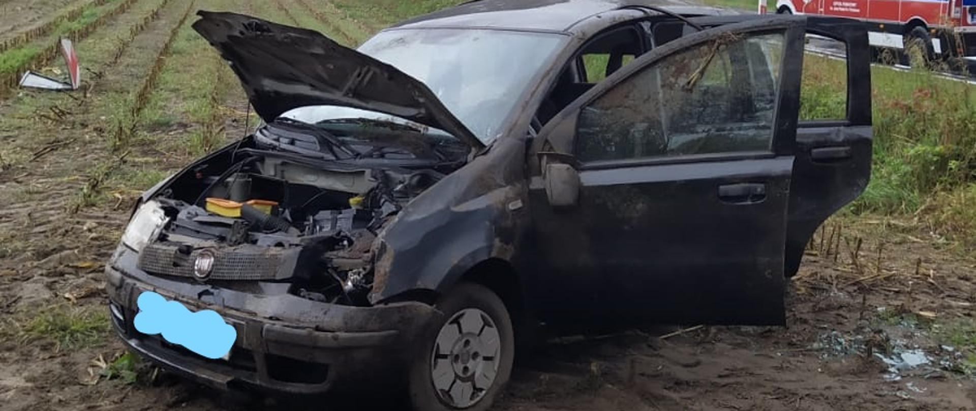 Rozbite auto po dachowaniu na polu. W tle pojazdy ratownicze.