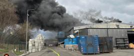 Na zdjęciu wykonanym z odległości kilkudziesięciu metrów znajduje się blaszana hala sortowni nad którą kłębi się czarny, gęsty dym. Na pierwszym planie, przed halą ustawione są kontenery obok, których rozciągnięta jest jedna z pierwszych linii wężowych.