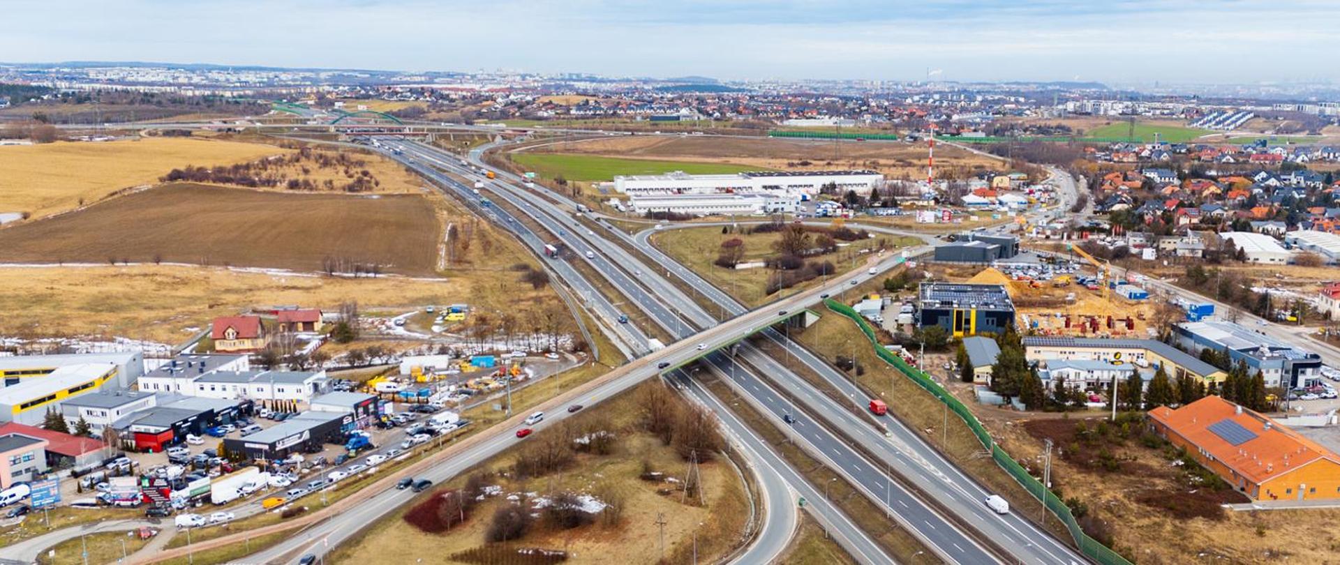 Panoramiczne ujęcie z lotu ptaka przedstawiające węzeł drogowy otoczony terenami zabudowanymi i polami. Kilka przecinających się dróg ekspresowych i lokalnych, mosty oraz zjazdy. Wokół widoczne budynki usługowe, magazyny, osiedla domów jednorodzinnych i tereny niezabudowane. W tle rozciąga się panorama miasta.
