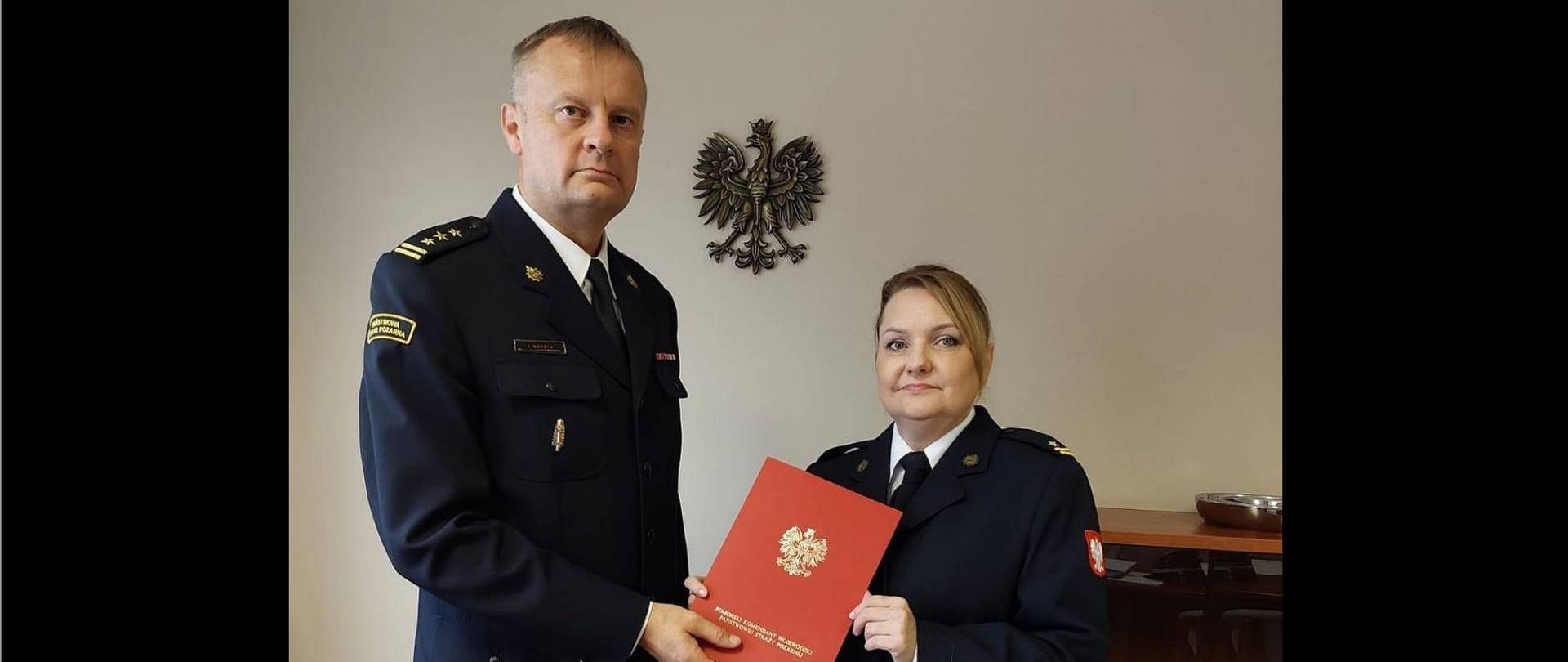 Zdjęcie przedstawia komendanta powiatowego wręczającego nagrodę wyróżnionej funkcjonariuszce. W tle godło polski.