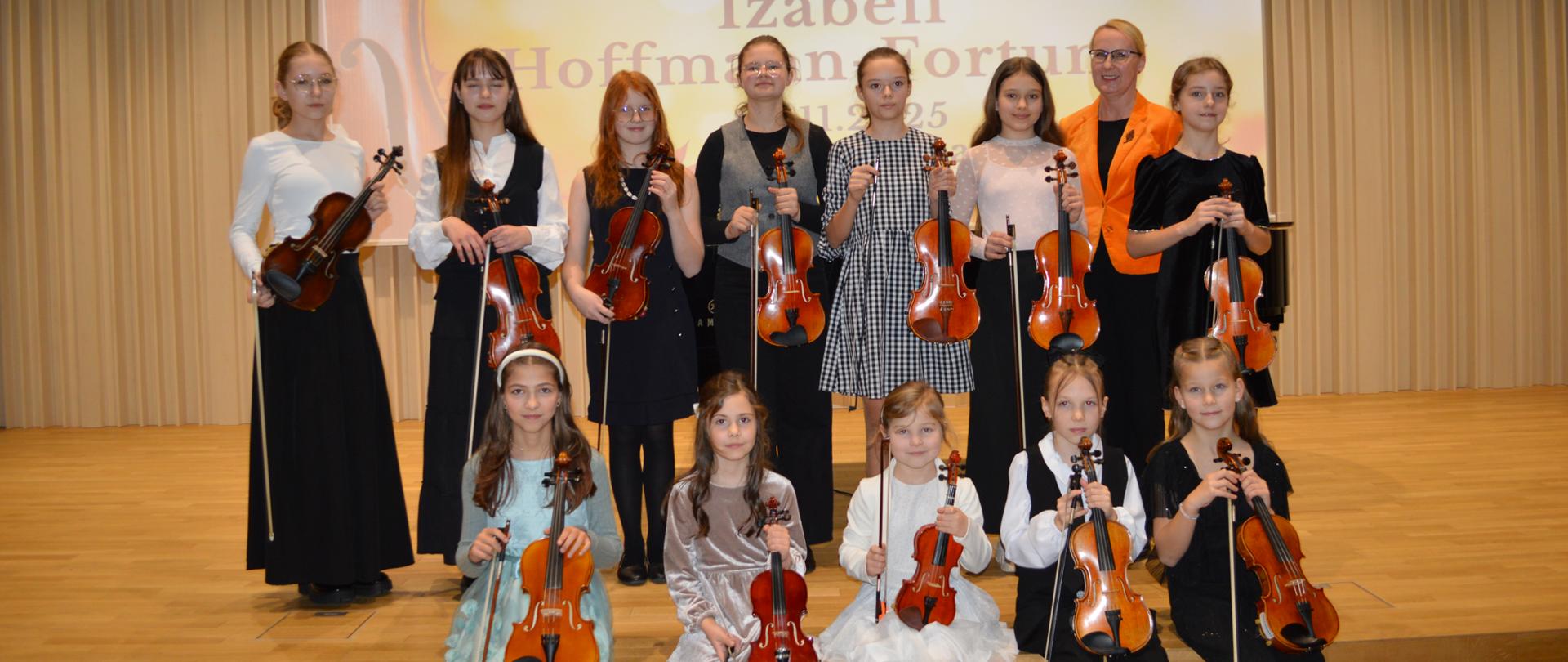 Na zdjęciu widoczna jest scena sali koncertowej z jasną, drewnianą podłogą i pionowymi panelami akustycznymi na ścianach. W tle znajduje się duży ekran projekcyjny z wyświetlonym plakatem koncertowym w jesiennej kolorystyce — dominują odcienie pomarańczu, złota i brązu, a po lewej stronie grafiki widoczny jest fragment skrzypiec oraz opadające liście klonu. Na plakacie czytelny jest napis „KONCERT klasy skrzypiec i absolwentek Izabeli Hoffmann-Fortuny”.
Na scenie ustawiona jest grupa młodych skrzypaczek – część stoi w rzędzie, a część siedzi na krawędzi podestu. Wszystkie trzymają skrzypce w dłoniach, niektóre także smyczki. Ubrane są w eleganckie stroje koncertowe: sukienki, spódnice i marynarki w stonowanych kolorach – czerni, bieli, beżu i szarości, z subtelnymi akcentami koronek i wzorów. Jedna z osób, stojąca blisko środka, wyróżnia się jaskrawym, pomarańczowym szalem lub elementem stroju, który nawiązuje do jesiennej tematyki plakatu.
Po prawej stronie sceny stoi czarny fortepian z zamkniętą klapą. Oświetlenie sufitowe tworzy ciepłą, przyjazną atmosferę. Cała grupa zwrócona jest przodem do obiektywu i pozuje do wspólnego zdjęcia po koncercie, z wyraźnie widocznym poczuciem dumy i radości z występu.