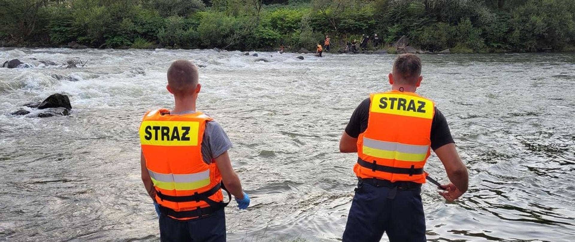 Na pierwszym planie dwóch strażaków stojących na brzegu rzeki ubranych w kamizelki asekuracyjne pomarańczowe z odblaskowym napisem "STRAŻ" na plecach oraz spodnie specjalne twarzami skierowanych w stronę rzeki. Na drugim planie na drugim brzegu rzeki strażacy prowadzą czynności ratownicze względem osoby przebywającej w wodzie.