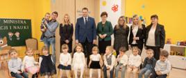 Zdjęcie zbiorowe, w sali z żółtymi ścianami na krzesełkach ustawionych w rzędzie siedzą małe dzieci, za nimi stoi kilkoro dorosłych, minister Czarnek pośrodku.