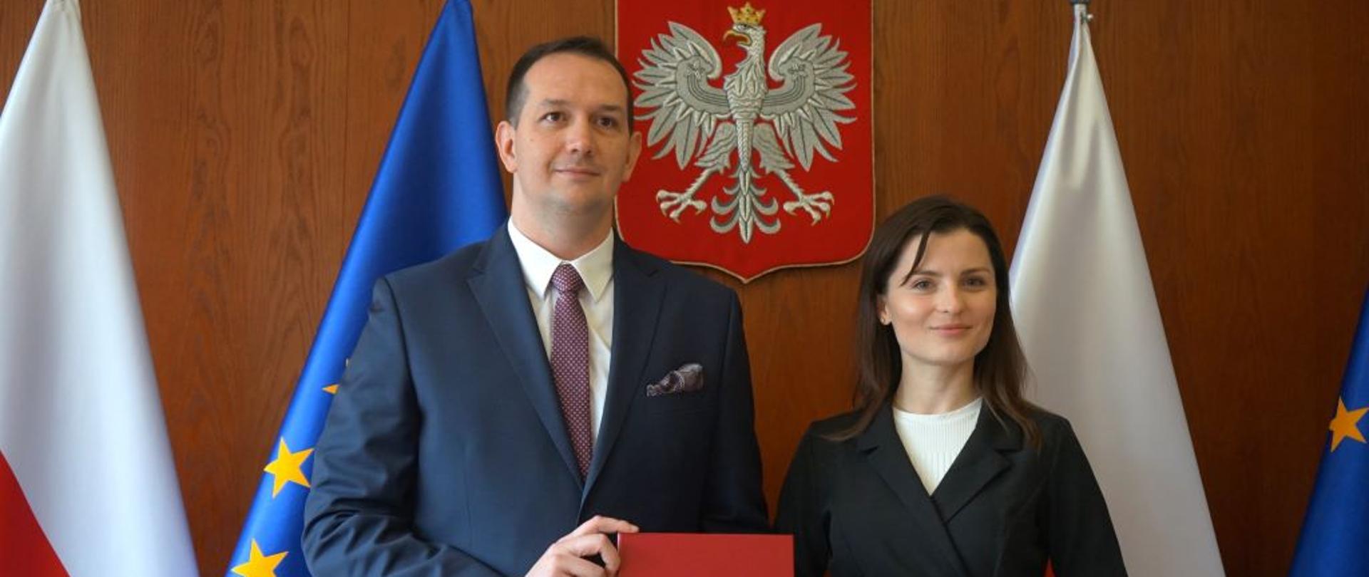 Mężczyzna w garniturze stoi obok kobiety i trzyma w ręku czerwoną teczkę z godłem narodowym. W tle flagi Polski i UE 