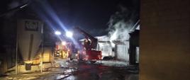 Rozstawiony podnośnik hydrauliczny w porze nocnej, wykorzystany podczas działań gaśniczych . Pożar budynku gospodarczego opanowany trwa dozorowanie miejsca pożaru. Miejsce zdarzenia jest oświetlone. Strażacy stoją przy samochodach ratowniczych obserwują ugaszony budynek .