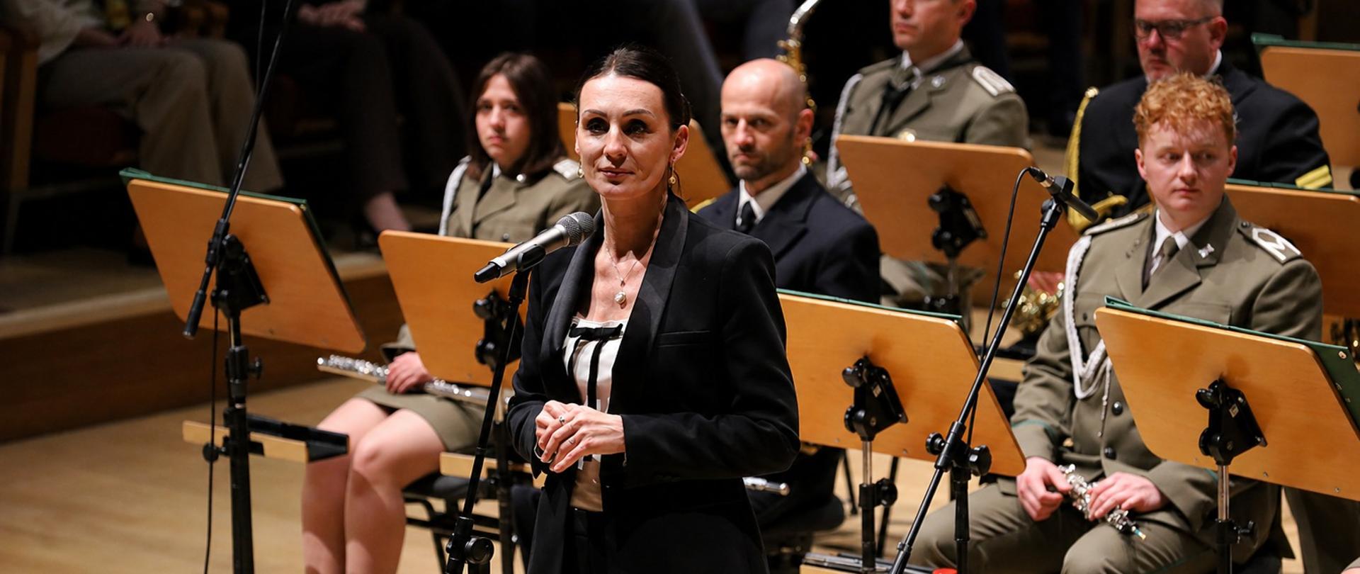 Kobieta stojąca przy mikrofonie na scenie sali koncertowej, ubrana elegancko w czarny garnitur, przemawia przed orkiestrą. Za nią siedzą muzycy w mundurach, trzymający instrumenty dęte, a przed nimi ustawione są pulpity z nutami. W tle widać publiczność siedzącą na widowni.
