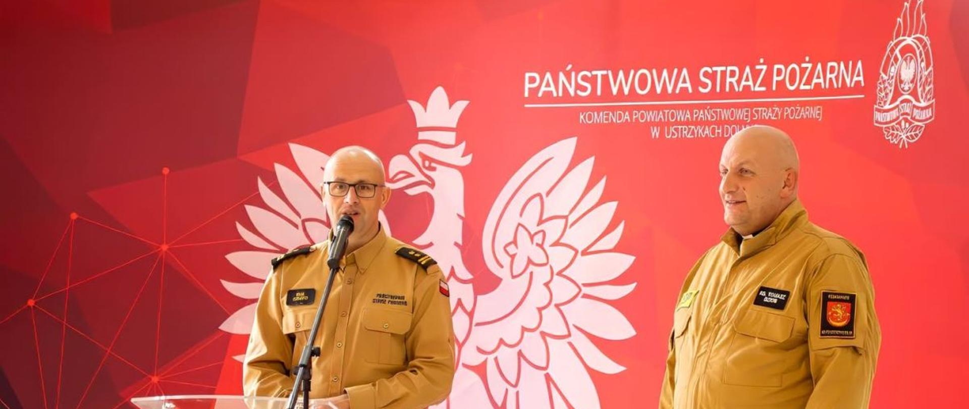 Zdjęcie przedstawia komendanta powiatowego PSP przy mównicy. Obok stoi kapelan strażaków powiatu bieszczadzkiego. W tle makieta z Orłem Białym i napisem oraz logiem Państwowej Straży Pożarnej. 