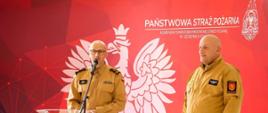 Zdjęcie przedstawia komendanta powiatowego PSP przy mównicy. Obok stoi kapelan strażaków powiatu bieszczadzkiego. W tle makieta z Orłem Białym i napisem oraz logiem Państwowej Straży Pożarnej. 