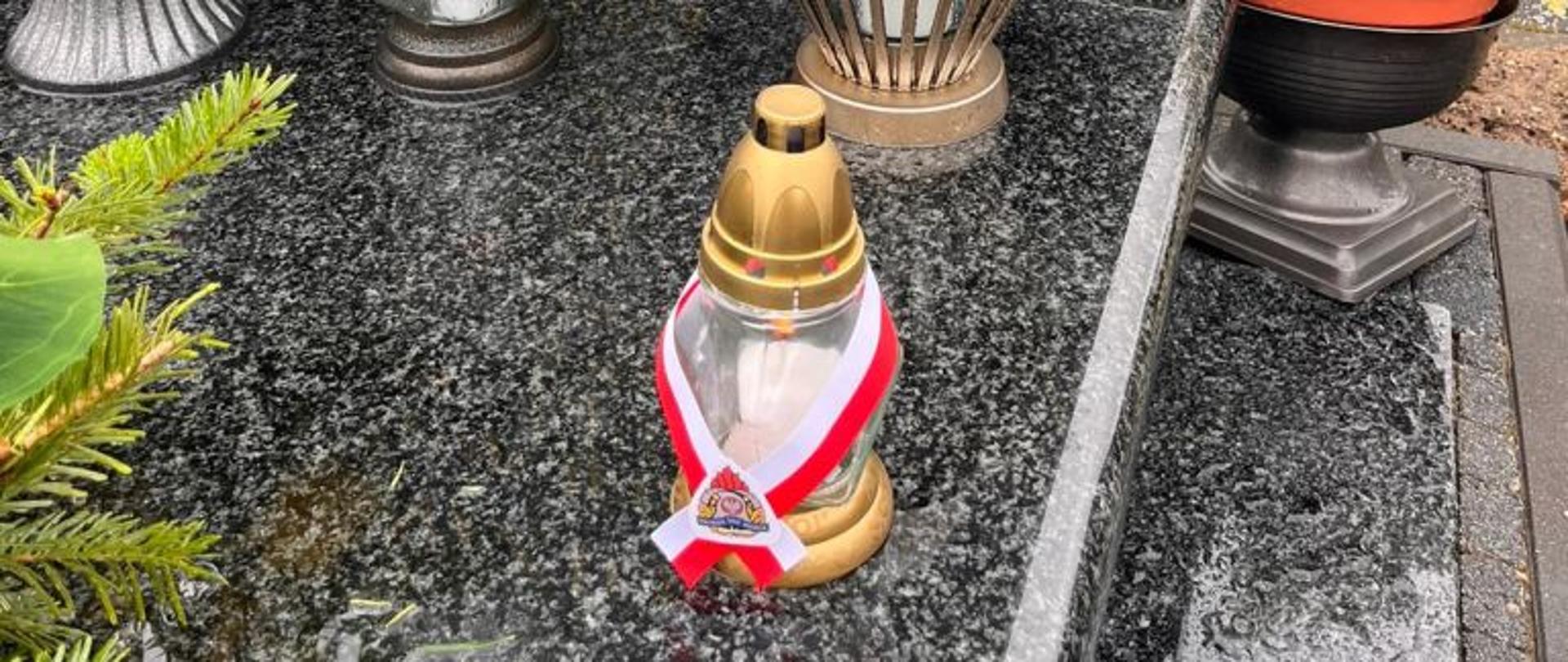 Centralny punkt zdjęcia stanowi nagrobek. Na płycie widoczna jest grafika przedstawiająca zmarłego strażaka. Na grobie kwiaty i znicze.