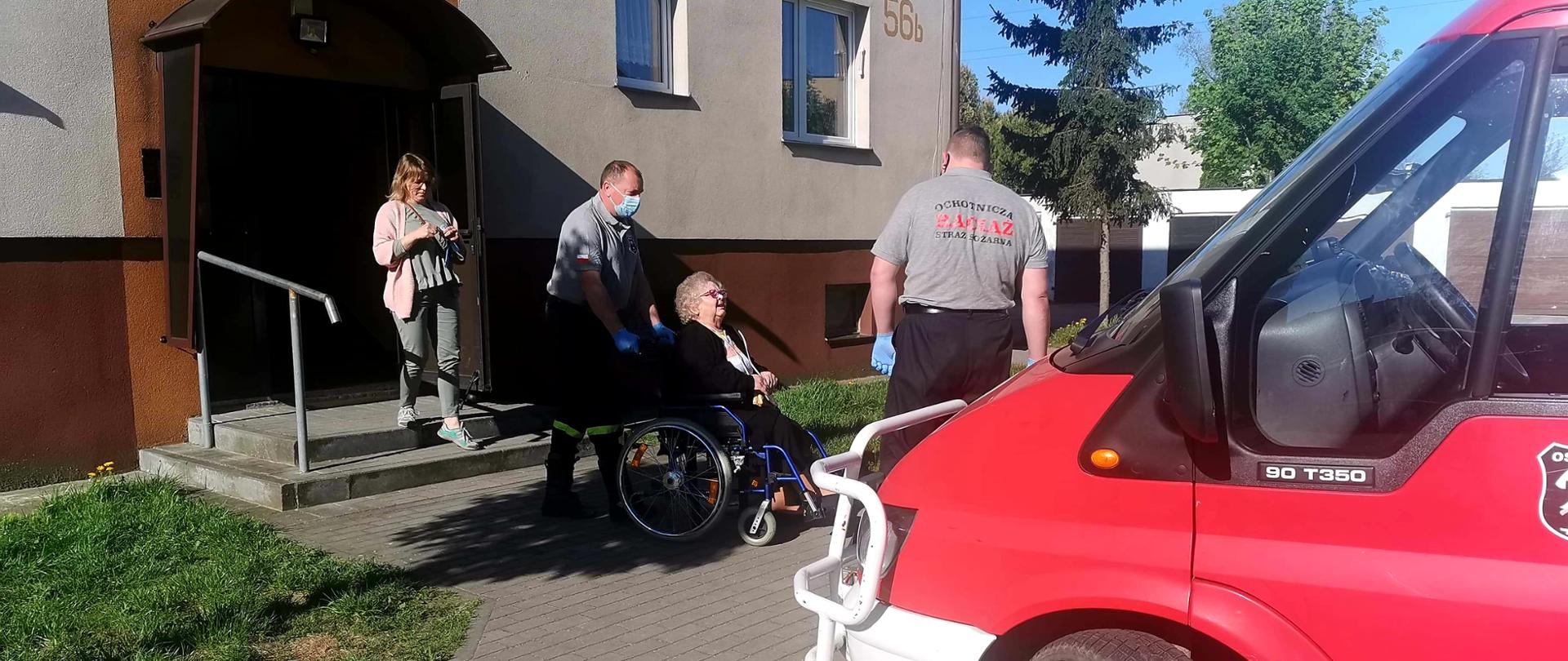 Starsza osoba na wózku inwalidzkim transportowana przez strażaka do samochodu pożarniczego. Z prawej strony stoi drugi strażak, przednik fragment samochodu pożarniczego. Z lewej strony budynek mieszkalny wielorodzinny i jedna osoba stojąca na schodkach.