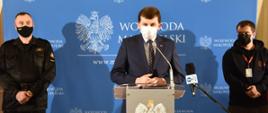 Na zdjęciu widoczni przy mównicy przemijający podczas briefingu prasowego dotyczącego przełączania numeru alarmowego 998 do Centrum Powiadamiania Ratunkowego wojewoda małopolski Łukasz Kmita i stojący po jego prawej stronie małopolski komendant wojewódzki Państwowej Straży Pożarnej st. bryg. Piotr Filipek a po lewej kierownik Centrum Powiadamiania Ratunkowego w Krakowie Łukasz Bombała. Briefing prasowy odbywał się w sali konferencyjnej Urzędu Wojewódzkiego w Krakowie.