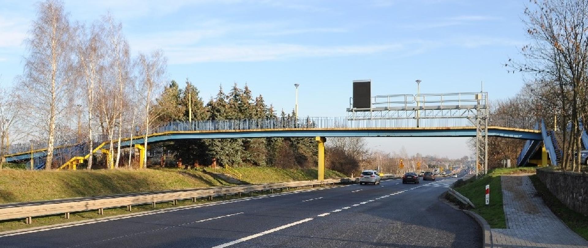 Kładka dla pieszych w ciągu DK92 w miejscowości Siedlec. Na zdjęciu widoczna droga nad którą znajduje się w pewnej odległości kładka dla pieszych. W oddali widoczne samochody. 