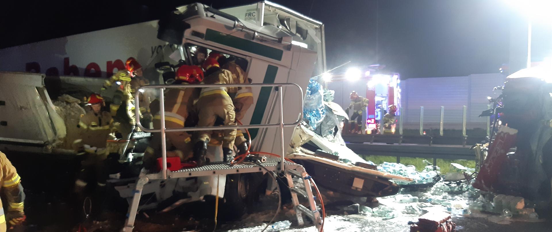 na zdjęciu samochód ciężarowy po wypadku, zniszczona kabina. Strażacy w ubraniach specjalnych na podeście ratowniczym wykonują dostęp do kierowcy znajdującego się w kabinie. Pora nocna.
