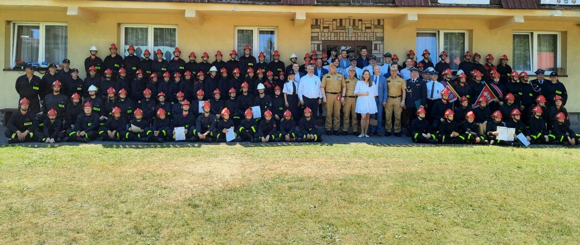 Przed żóltym budynkiem z brązowym dachem, grupowe zdjęcie członków Młodzieżowych Drużyn Pożarniczych ubranych w czarne mundury koszarowe uczestników obozu, ustawionych w czterech rzędach, kadry obozu oraz zaproszonych gości.