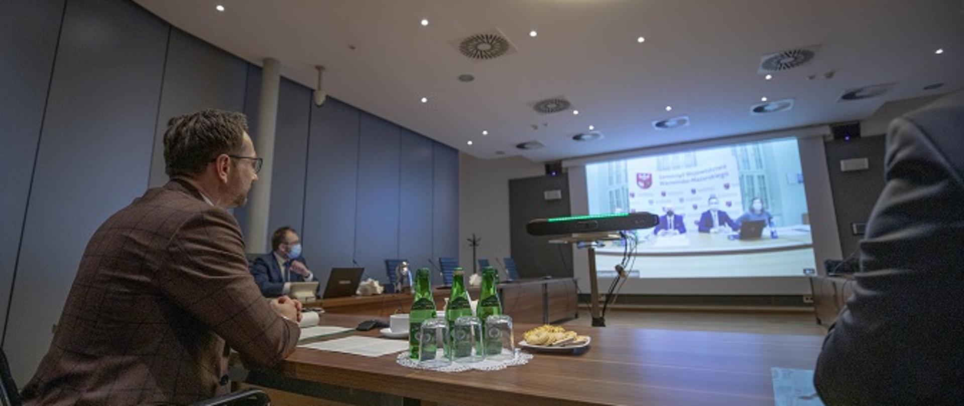 Na zdjęciu widać wiceministra Waldemara Budę siedzącego przy stole konferencyjnym na przeciwko wielkiego ekranu, na którym transmitowana się wideokonferencja z marszałkiem woj. warmińsko-mazurskiego.