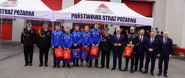 Młodzieżowa Drużyna Pożarnicza Ochotniczej Straży Pożarnej w Szadkowicach wraz z trenerem, Zastępcą Komendanta Wojewódzkiego Państwowej Straży Pożarnej w Łodzi, Zastępcą Komendanta Wojewódzkiego Państwowej Straży Pożarnej w Opocznie oraz z zaproszonymi gośćmi stoją przed namiotami z napisem „Państwowa Straż Pożarna”. Członkowie drużyny w niebieskich bluzach prezentują medale, trzymają torby z nagrodami otrzymanymi podczas uroczystości. 