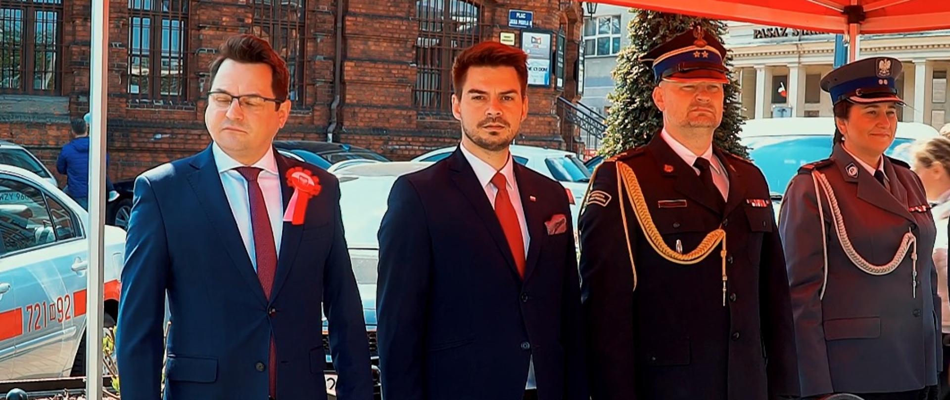 Komendant powiatowy PSP w Żyrardowie st. bryg. Tomasz Cybul stoi wśród zaproszonych gości pod czerwonym namiotem. Po jego lewej stronie stoi komendant powiatowy policji, po jego lewej stronie przewodniczący rady powiatu żyrardowskiego i jeszcze jeden mężczyzna.