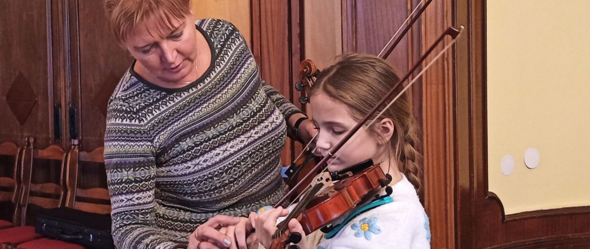 Zdjęcie kolorowe. Dziewczynka ze skrzypcami pracuje pod kierunkiem jasnowłosej kobiety we wzorzystym swetrze. W tle zabytkowe, dwuskrzydłowe drzwi, krzesła i czarny skrzypcowy futerał.