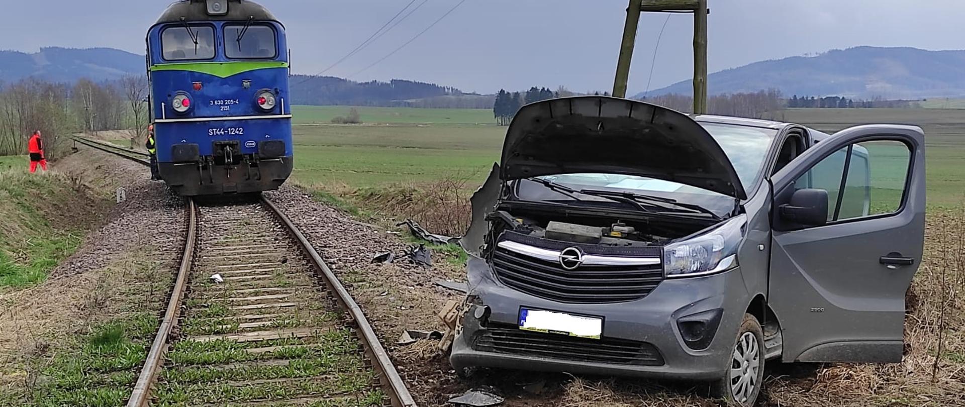 Pociąg w kolorze niebieskim ustawiony na torach, po prawej stronie bliżej oglądającego fotografię widoczny samochód osobowy, bus koloru grafitowego drzwi pojazdu otwarte od prawej strony. Pojazd osobowy ustawiony w kierunku patrzącego. Zdjęcie wykonano na terenie niezurbanizowanym, widoczne tereny zielone.