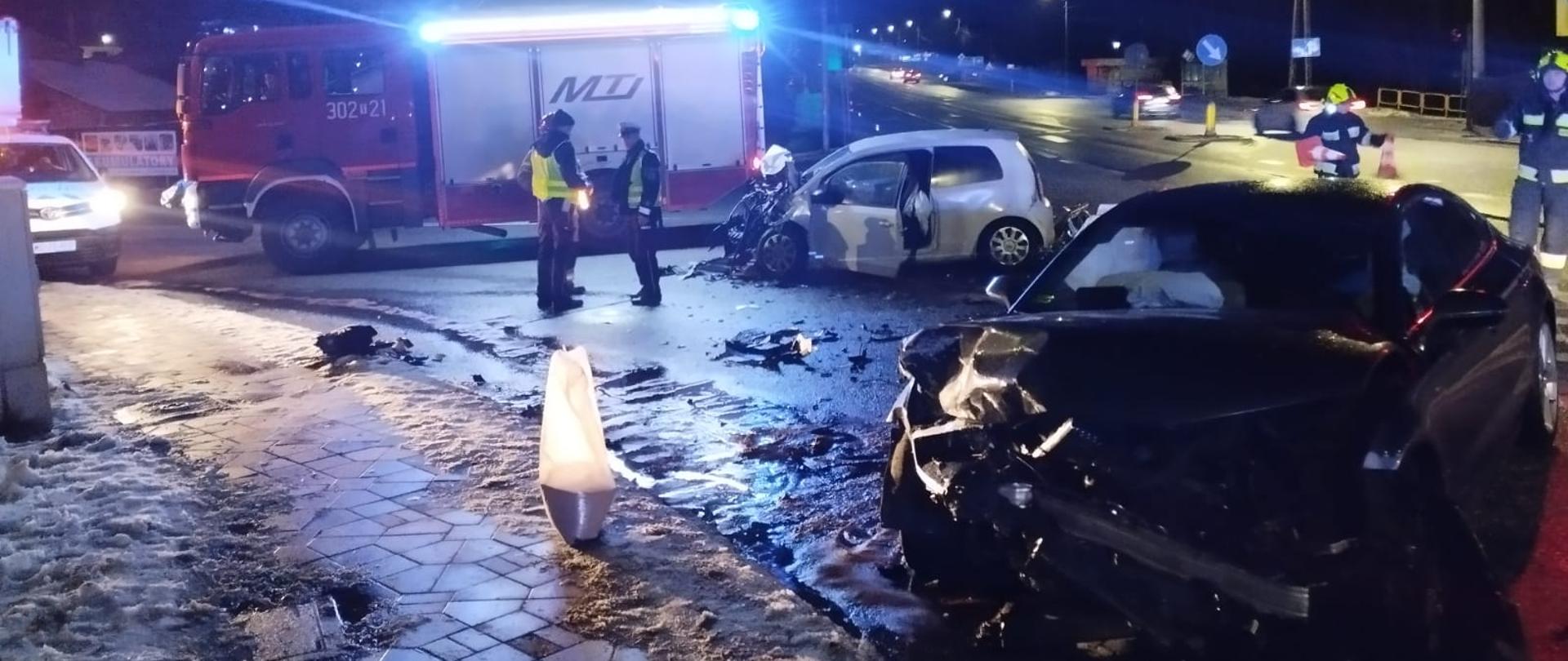 Zdjęcie przedstawia dwa zniszczone w wyniku zderzenia auta osobowe. W tle stoi samochód pożarniczy na którego tle stoją dwaj funkcjonariusze Policji.