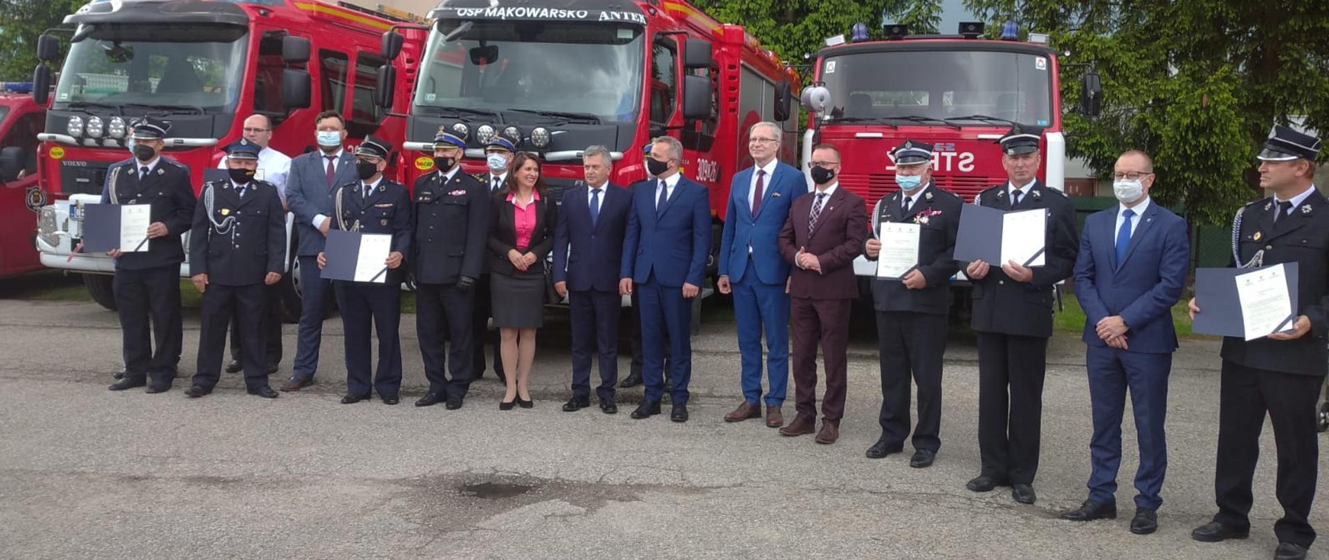 Na zdjęciu kilkunastu druhów OSP w mundurach wyjściowych koloru granatowego wraz z obecnymi na uroczystości parlamentarzystami, wojewodą oraz przedstawicielami PSP. Wszyscy stoją na placu przy remizie OSP Koronowo, za nimi w tle 3 samochody pożarnicze ciężarowe koloru czerwonego stojące przodem do osoby robiącej zdjęcie.