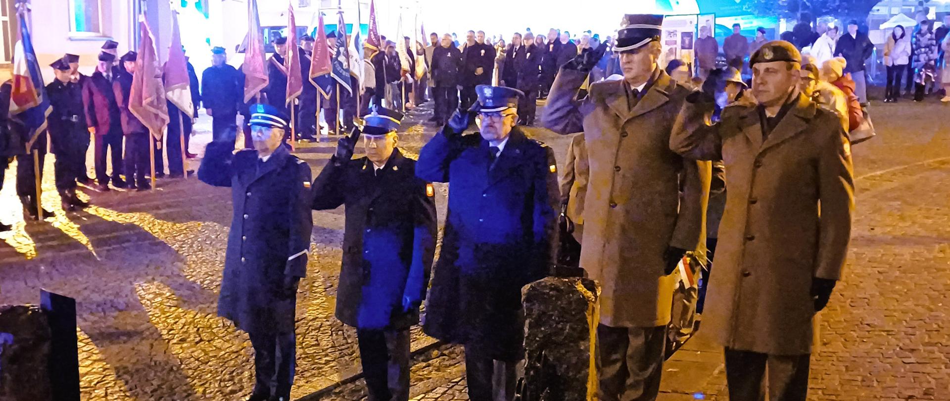 Uroczystość złożenia kwiatów przez przedstawicieli służb mundurowych pod tablicą upamiętniającą 100. rocznicę odzyskania niepodległości przez Polskę. Na pierwszym planie pięciu mężczyzn w mundurach stoi na baczność i salutuje, za nimi widoczny jest szpaler pocztów sztandarowych oraz zgromadzeni uczestnicy uroczystości w wieczornym oświetleniu miejskim.