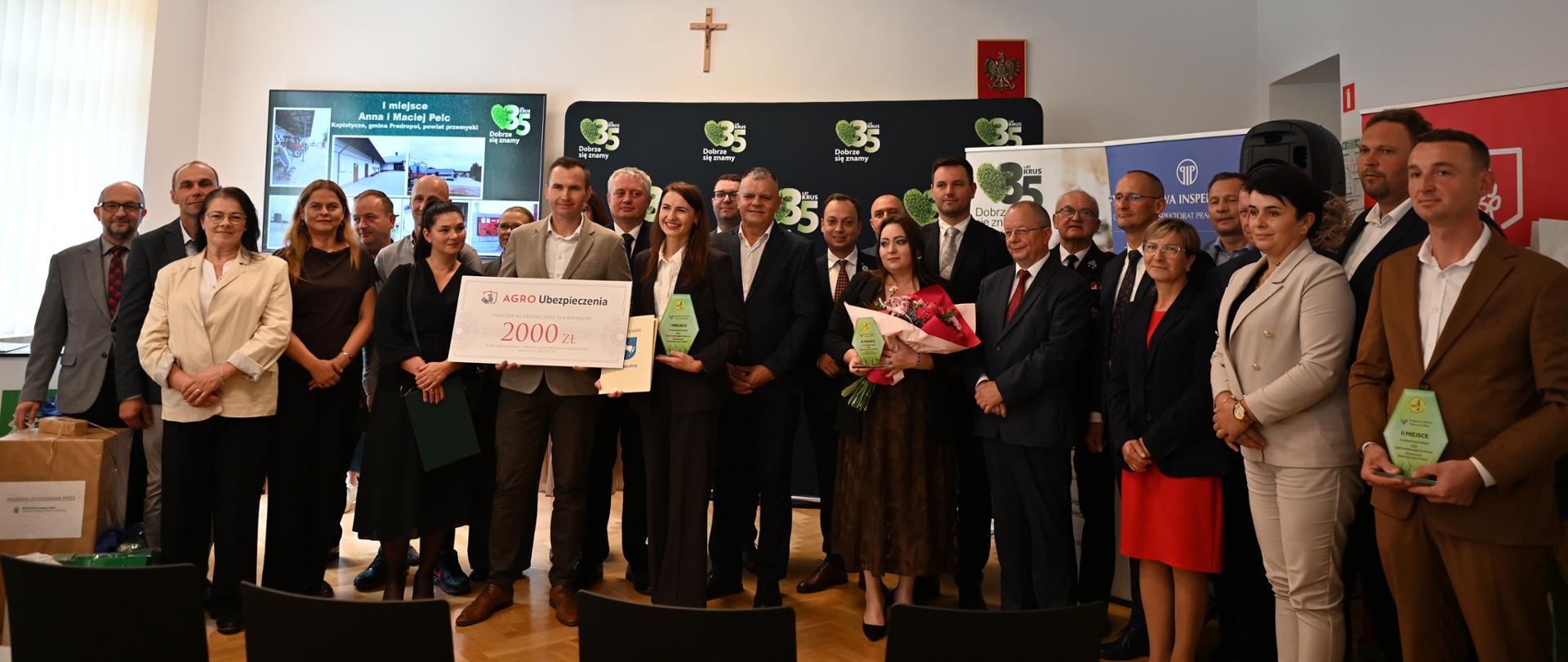 Kilkadziesiąt osób pozuje do zdjęcia na tle ścianki z logo 35-lecia KRUS. Dwie kobiety i mężczyzna trzymają w rękach zielone statuetki z logo konkursu BGR, a jeden z mężczyzn trzyma symboliczny czek na 2000 zł. Wszyscy uśmiechają się do zdjęcia.