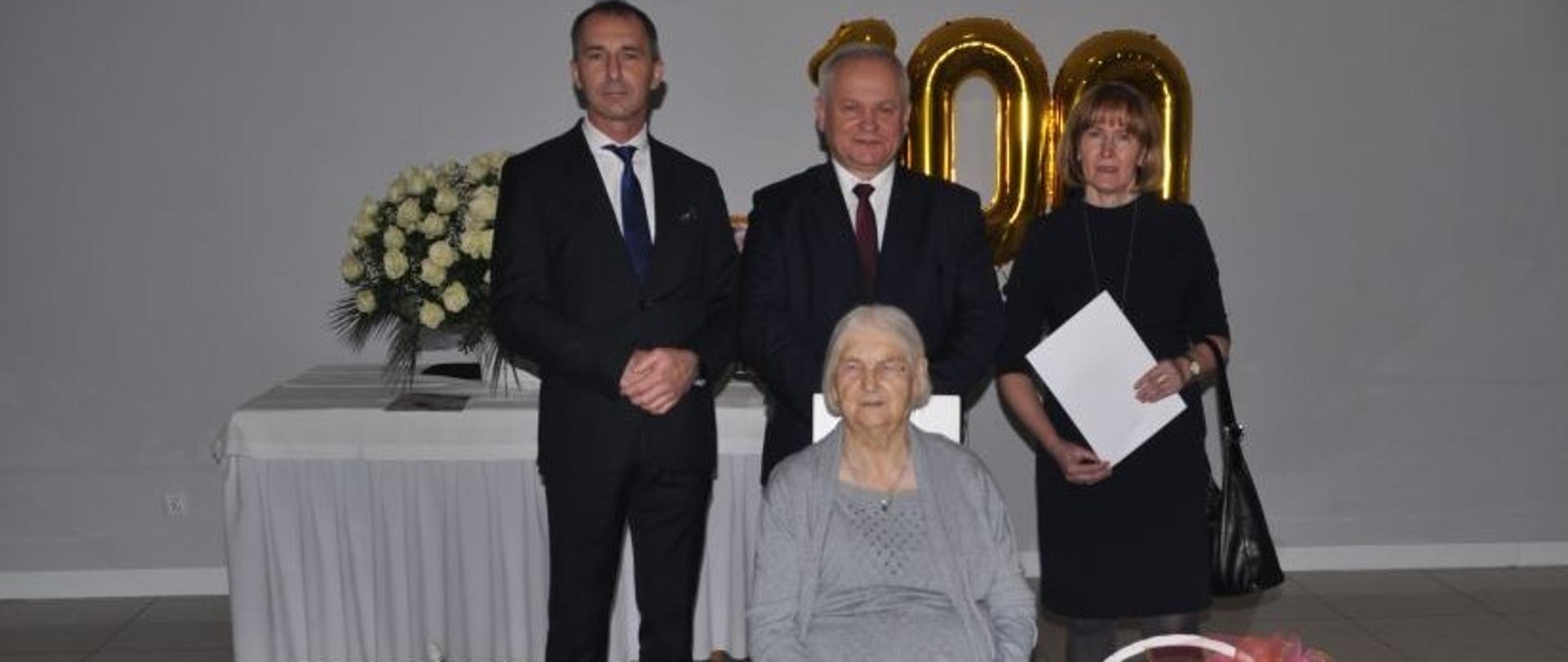 Starsza kobieta siedzi na krześle lekko się uśmiechając. W dłoniach trzyma dyplomy a wokół niej stoją kosze z kwiatami i ozdobne torby z prezentami. Z tyłu stoją dwaj mężczyźni i kobieta. Za nimi stół z bukietem białych róż i złotymi balonami w kształcie liczby 100.