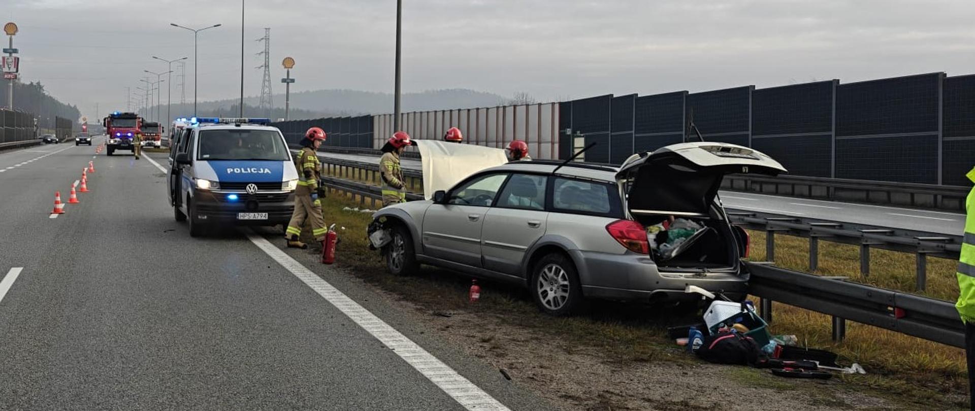 Zdjęcie przedstawia miejsce wypadku: z prawej strony, na poboczu stoi uszkodzony samochód, za nim widać stojący radiowóz , a w głębi 2 pojazd strażackie. Przy zniszczonym aucie pracują strażacy.