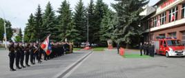 Zdjęcie przedstawia uroczystą zbiórkę funkcjonariuszy z okazji Dnia Flagi przed Jednostką Ratowniczo-Gaśniczą nr 1 w Rzeszowie. Z lewej strony ustawiony jest pododdział funkcjonariuszy wraz z pocztem sztandarowym i flagowym. Z prawej strony znajdują się komendanci miejscy oraz wojewódzcy PSP w Rzeszowie obok pojazdu specjalnego.