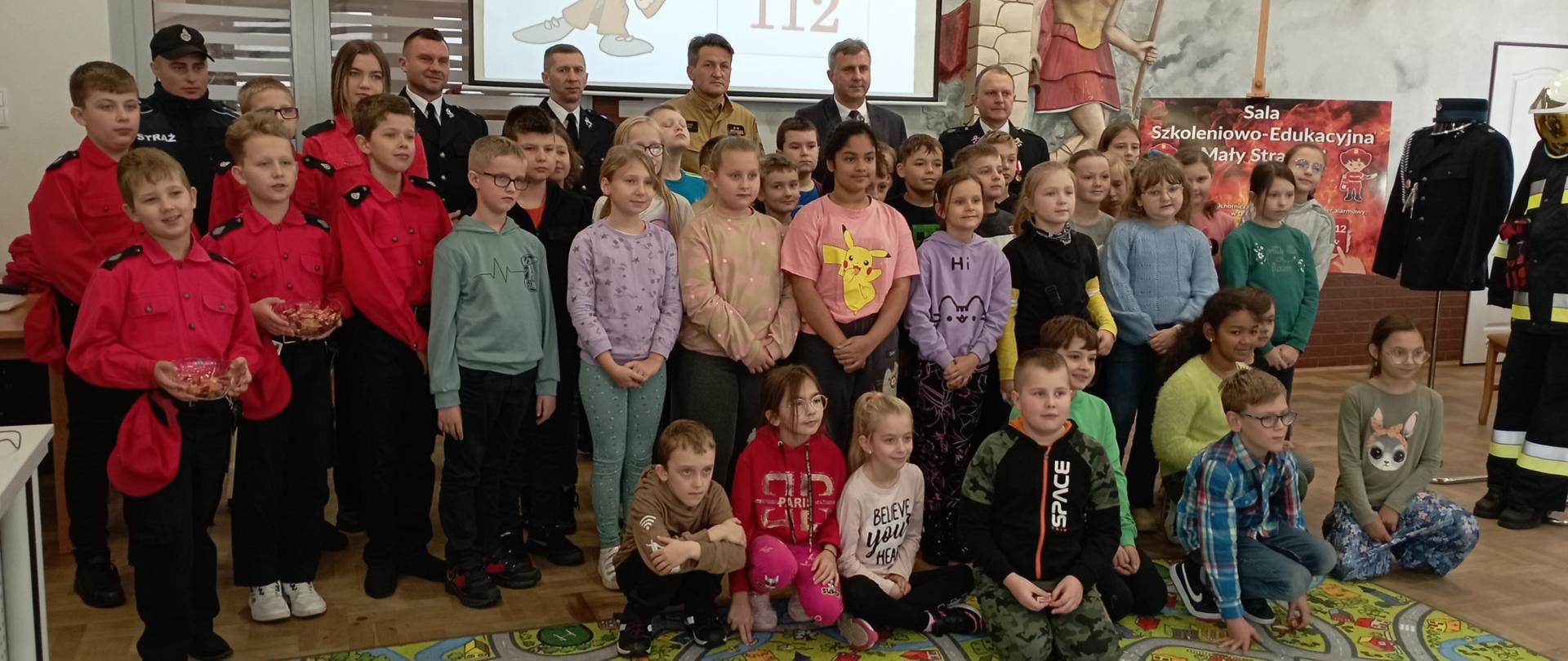 Zdjęcie przedstawia uczestników pierwszych zajęć w salce edukacyjnej „Mały strażak” wraz z organizatorami i zaproszonymi gośćmi.