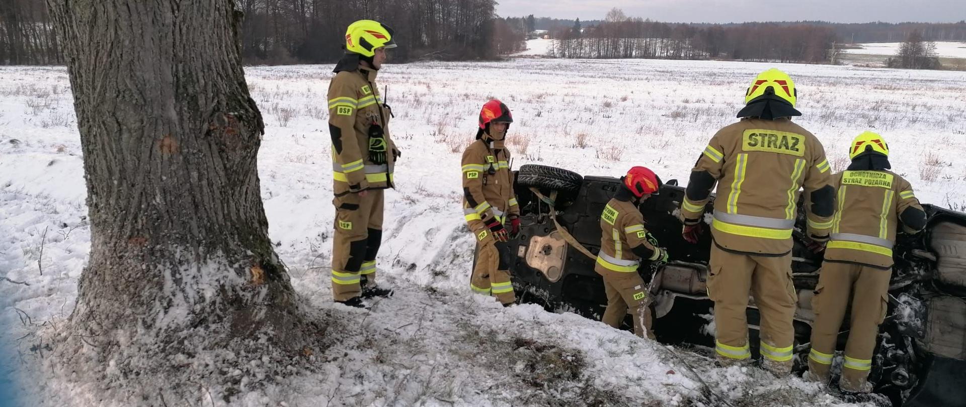Zima, dużo śniegu. W rowie na poboczu drogi leży auto osobowe na boku. Przy aucie strażacy w jasnych mundurach i hełmach. Z lewej strony stoi drzewo.
