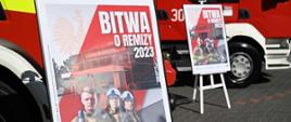 Zdjęcie przedstawia dwa plakaty akcji "Bitwa o Remizy 2023". W tle samochód pożarniczy.