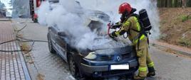 Pożar samochodu osobowego. Ulica Englerta w Rawiczu. Na jezdni stoi samochód osobowy koloru ciemnego spowity białym dymem. W tle fragment pojazdu straży pożarnej. Z przodu płonącego auta znajduje się strażak zabezpieczony sprzętem ochrony układu oddechowego, który gasi pożar. Auto posiada uszkodzona szybę czołową i przednią część. Z lewej strony, w oddali na chodniku stoi grupka ludzi. Z prawej strony - lasek miejski.