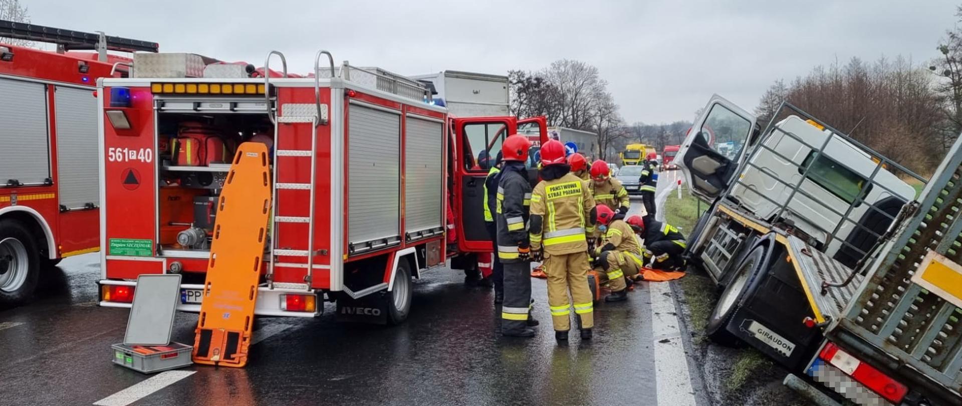 na drodze ustawione są samochody straży pożarnej w przydrożnym rowie leży samochód ciężarowy a pomiędzy tymi samochodami stoją strażacy, którzy obsługują niezbędny do działań ratowniczo-gaśniczych sprzęt. 