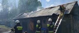 Budynek gospodarczy po pożarze. Trwa dogasanie. Przy budynku pracujący strażacy, jeden z nich pracuje na drabinie.