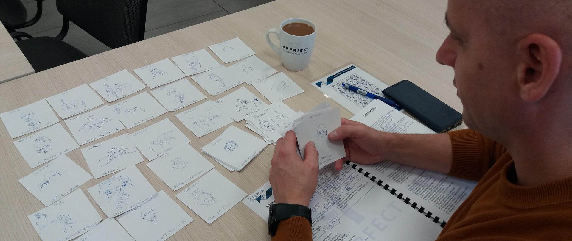 Zdjęcie przedstawia kartki - fiszki - wyłożone na stole. Mężczyzna przypomina sobie, jak pokazywać poszczególne gesty w ramach języka migowego.