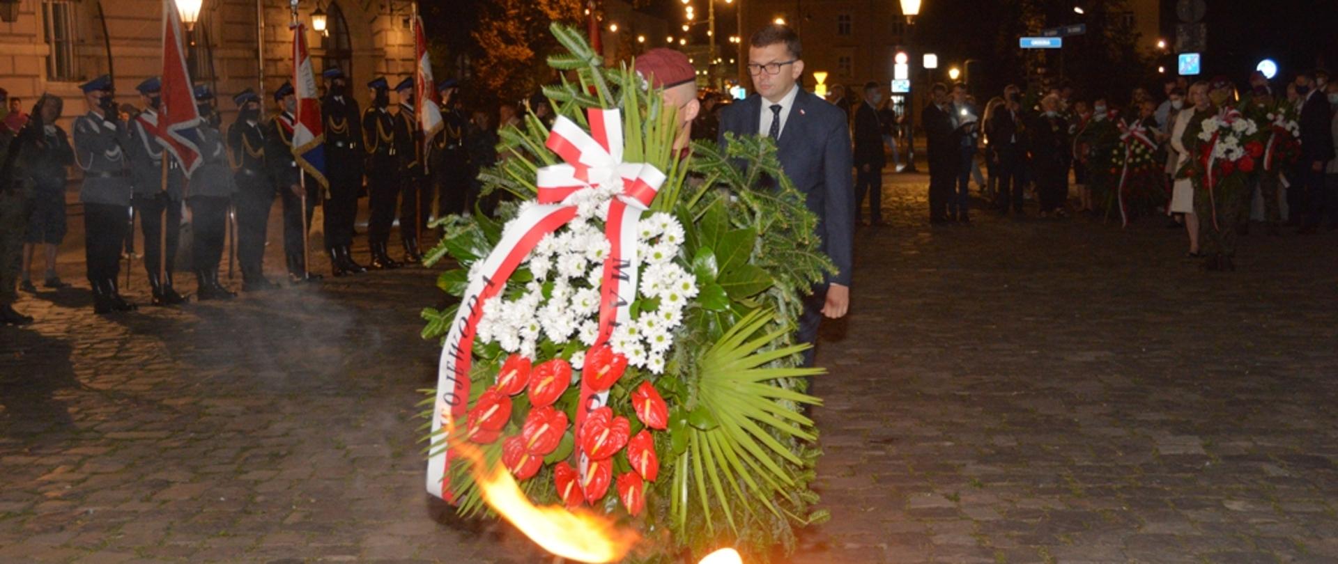 Zdjęcie zrobione nocą . Przedstawia delegację składającą wieniec. W tle ustawione są poczty sztandarowe.