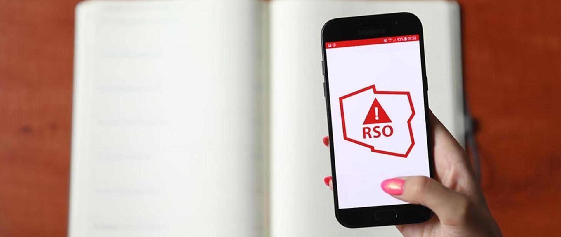 Zdjęcie przedstawia dłoń kobiety z pomalowanymi na różowo paznokciami, trzymającą czarny smartfon na tle otwartego, pustego notesu. Na ekranie telefonu widoczne jest białe tło z czerwonym logo aplikacji RSO (Regionalny System Ostrzegania), przedstawiającym zarys mapy Polski z wpisanym w środek trójkątnym znakiem ostrzegawczym i wykrzyknikiem.