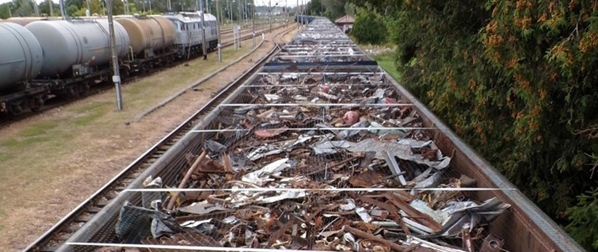 Wagon kolejowy wypełniony różnorodnymi odpadami metalowymi i przemysłowymi, m.in. zardzewiałe blachy, fragmenty maszyn, kable, zużyte części samochodowe oraz elementy z tworzyw sztucznych.